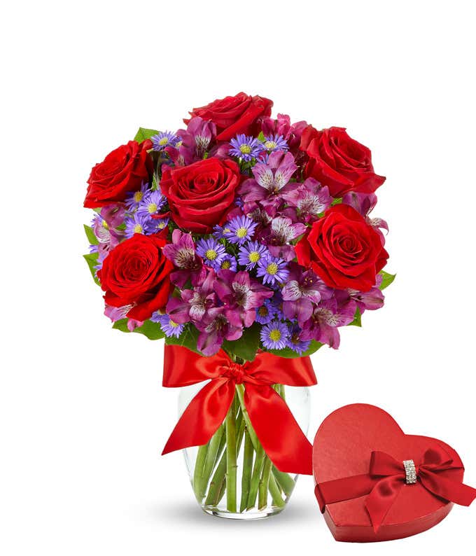 Bouquet of red roses with lots of smaller purple flowers in a clear glass vase with a red bow next to a box of chocoaltes
