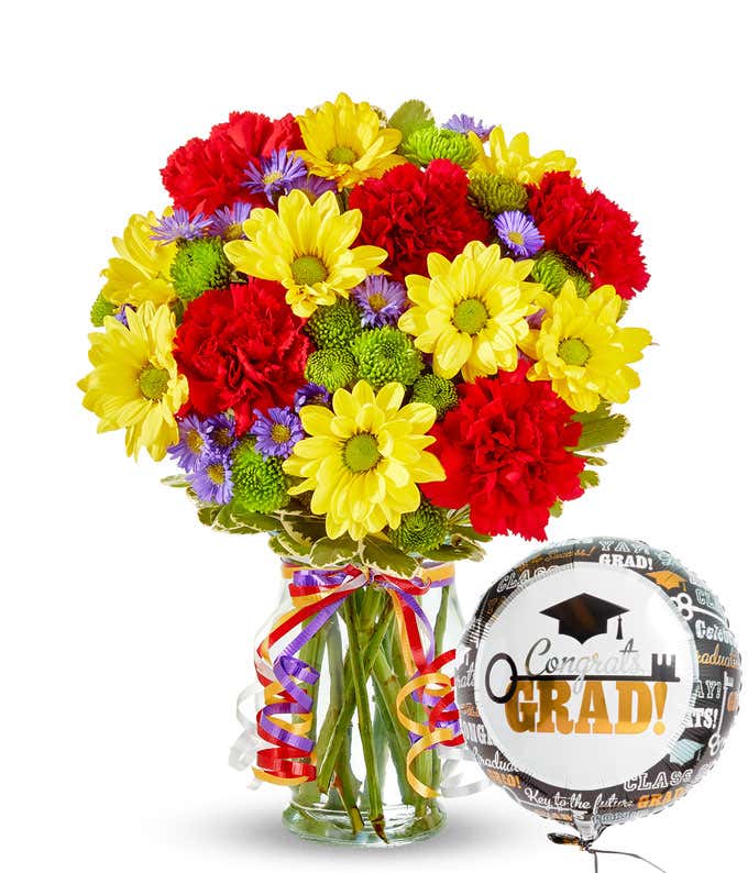 Bouquet of red carnations, yellow daisies, and green and purple filler flowers in a clear vase with colorful curled ribbons and a 'congrats grad!' mylar balloon. 