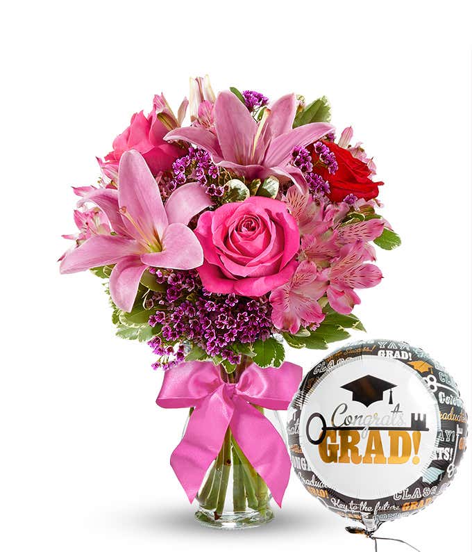 A bursting full bouquet of red and pink flowers including roses, lilies, and more. Next to a congratulations mylar balloon. 
