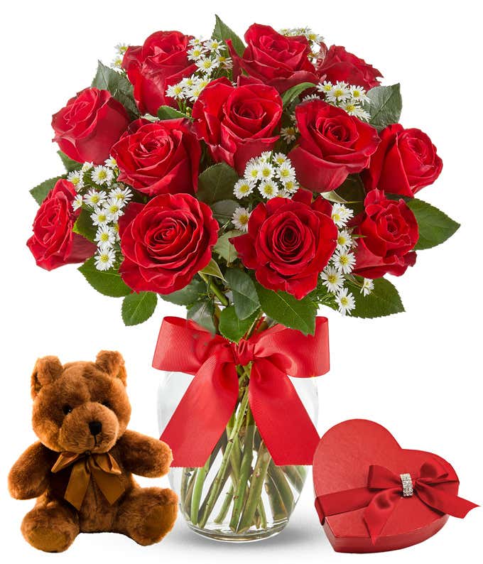 One dozen red roses with small white flowers and green leaves in a clear vase tied with a red bow. Next to a box of chocolates and a light brown teddy bear.