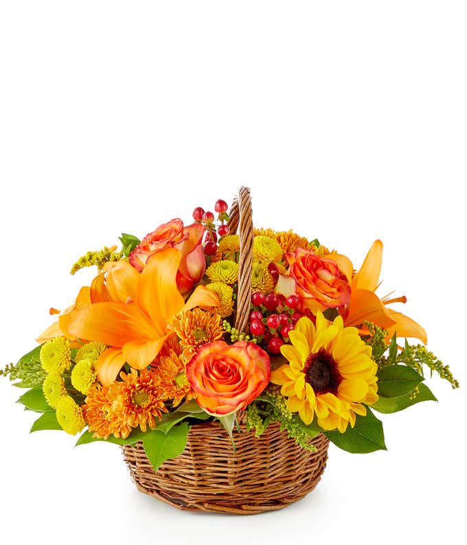 A vibrant floral arrangement featuring yellow Asiatic lilies, deluxe size orange roses, regular size orange carnations, yellow button poms, bronze cushion poms, red hypericum, and solidago, all beautifully presented in a basket.