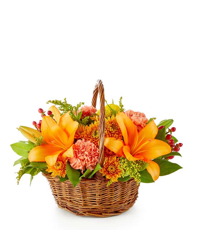 A vibrant floral arrangement featuring yellow Asiatic lilies, deluxe size orange roses, regular size orange carnations, yellow button poms, bronze cushion poms, red hypericum, and solidago, all beautifully presented in a basket.