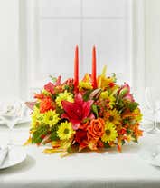 Alternative image 1 for A floral centerpiece of deep red lilies and bright orange roses accented by yellow daisies, red berries, and seeded eucalyptus formed into an oval centerpiece with 2 tall orange tapered candles