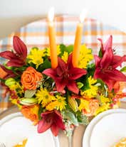 Alternative image 2 for A floral centerpiece of deep red lilies and bright orange roses accented by yellow daisies, red berries, and seeded eucalyptus formed into an oval centerpiece with 2 tall orange tapered candles
