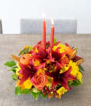Alternative image 3 for A floral centerpiece of deep red lilies and bright orange roses accented by yellow daisies, red berries, and seeded eucalyptus formed into an oval centerpiece with 2 tall orange tapered candles