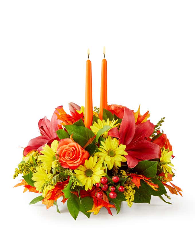 A floral centerpiece of deep red lilies and bright orange roses accented by yellow daisies, red berries, and seeded eucalyptus formed into an oval centerpiece with 2 tall orange tapered candles