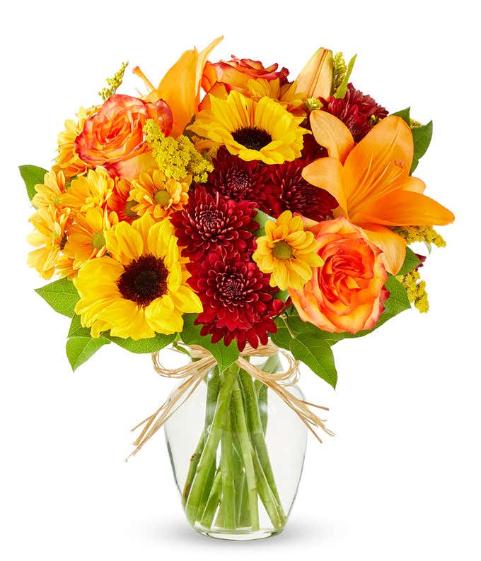 A vibrant fall bouquet in clear glass vase, sunflowers and orange lilies and roses are surrounded by yellow and maroon filler flowers in a clear glass vase.