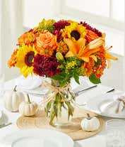 Alternative image 1 for A vibrant fall bouquet in clear glass vase, sunflowers and orange lilies and roses are surrounded by yellow and maroon filler flowers in a clear glass vase.