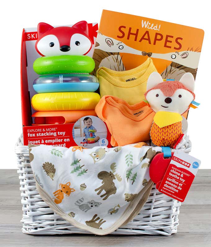 A white basket containing a fox stacking toy, a colorful onesie, a cozy blanket, a plush fox toy, and a book titled 'Wild Shapes'.