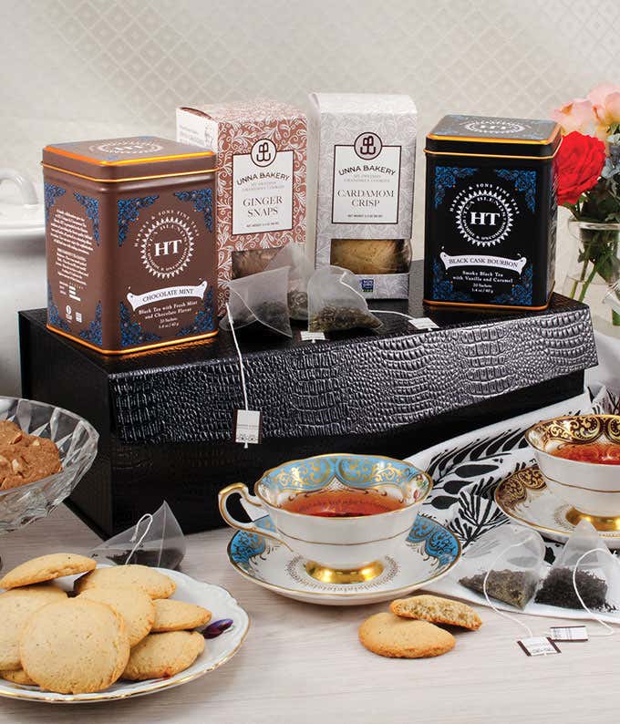 Elegant tea gift set featuring tins of Harney & Sons teas with two ornate teacups filled with tea, surrounded by assorted cookies, packaged tea bags, and a floral arrangement.