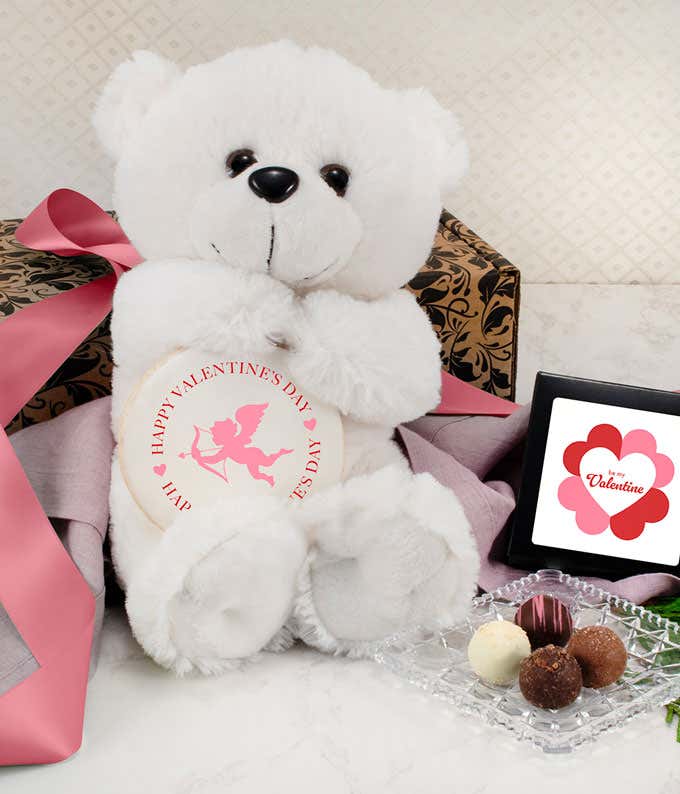 Valentine's Day gift set featuring a white plush teddy bear holding a round sign with a cupid design, a decorative box tied with a pink ribbon, a small framed card with a heart-shaped flower design, and a glass dish displaying assorted chocolate truffles.