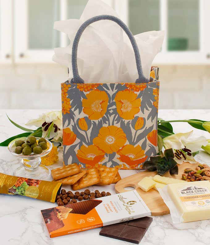 Floral-patterned gift tote filled with gourmet snacks including crackers, cheese, olives, chocolate, and nuts, arranged on a marble surface with white lilies and greenery.