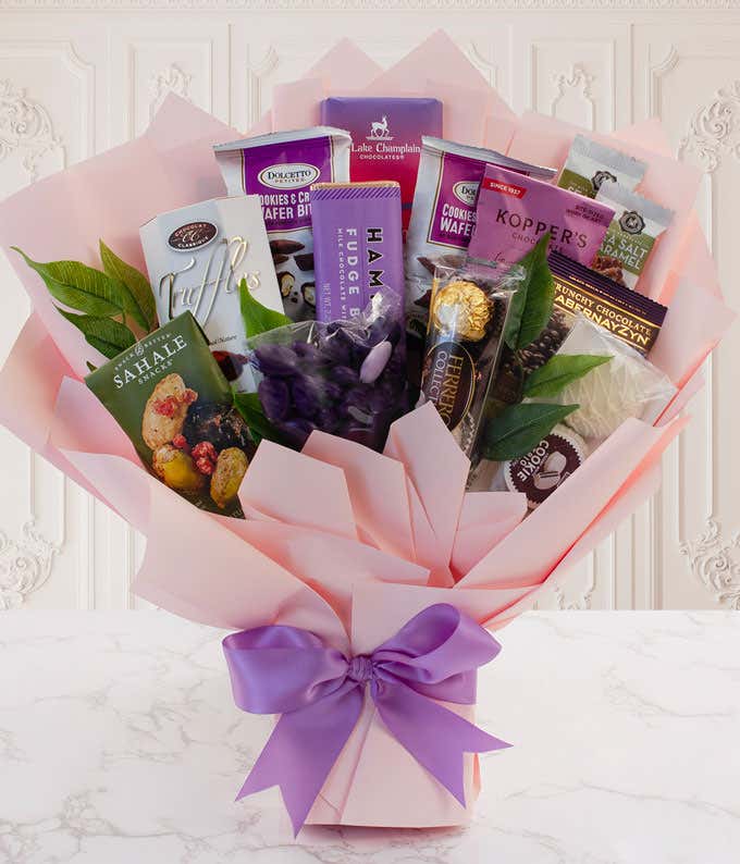 Pink gift bouquet filled with assorted gourmet chocolates, cookies, truffles, and snacks, arranged with greenery and wrapped in blush paper with a purple ribbon bow.