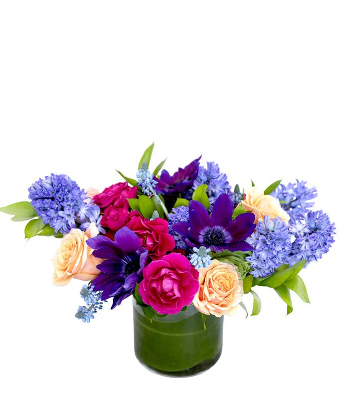 Purple, pink, and peach flowers arranged into a short leaf-lined cylinder vase