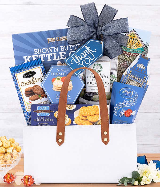 This elegant Thank You gift basket features a white tote with leather handles and a blue bow. It's filled with Tom Clark's Kettle Corn, Lindt Lindor Truffles, Vino e Formaggio Cheese Spread, Bella Campagna Olives, Ciokini cookies, and crackers. ​