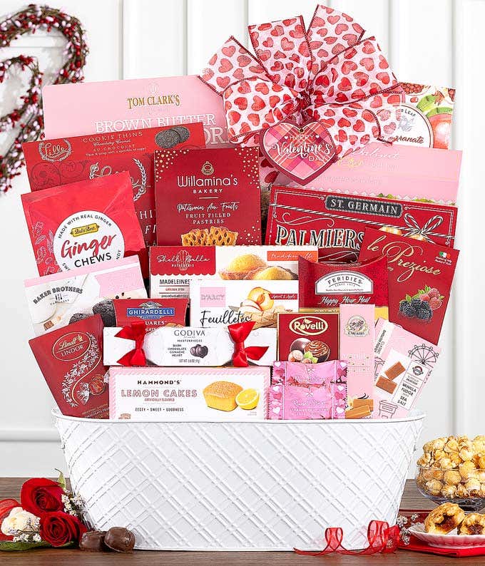 White gift basket filled with assorted gourmet treats in red and pink packaging, including chocolates, cookies, pastries, and popcorn, adorned with a red and pink heart-patterned ribbon.