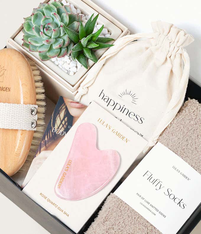 Top-down view of a self-care gift box featuring a potted succulent arrangement, dry body brush, rose quartz gua sha, fluffy socks, and a cotton drawstring pouch labeled 