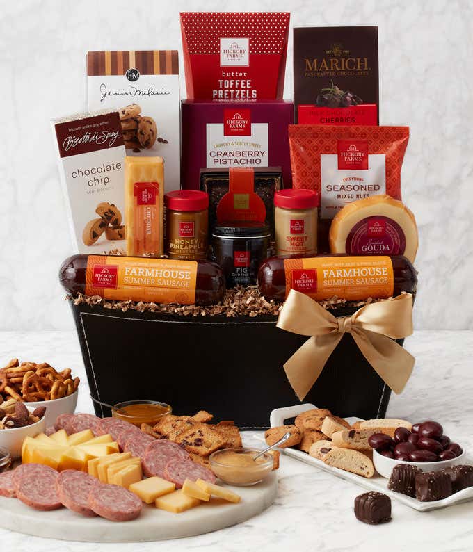 A  black gift basket with two packs of sausage, a wheel of gouda cheese, a block of cheddar cheese, a jar of honey pineapple mustard, a jar of hot & sweet mustard, and fig chutney, a box of chocolate chip mini biscotti, chocolate chip cookies, toffee pret