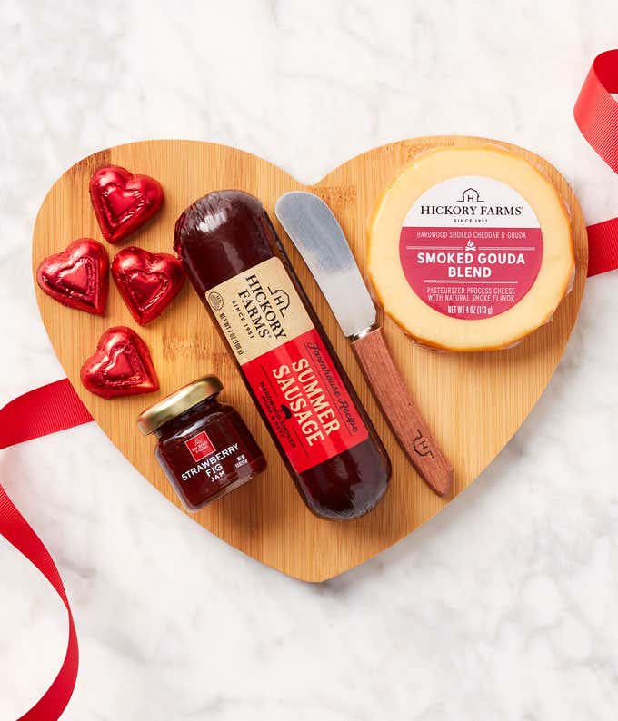 A serving board with a stick of salami, block of cheddar, boxes of cranberry pistachio crisps and english toffee, cheese spreader, and foil-wrapped chocolate hearts. 