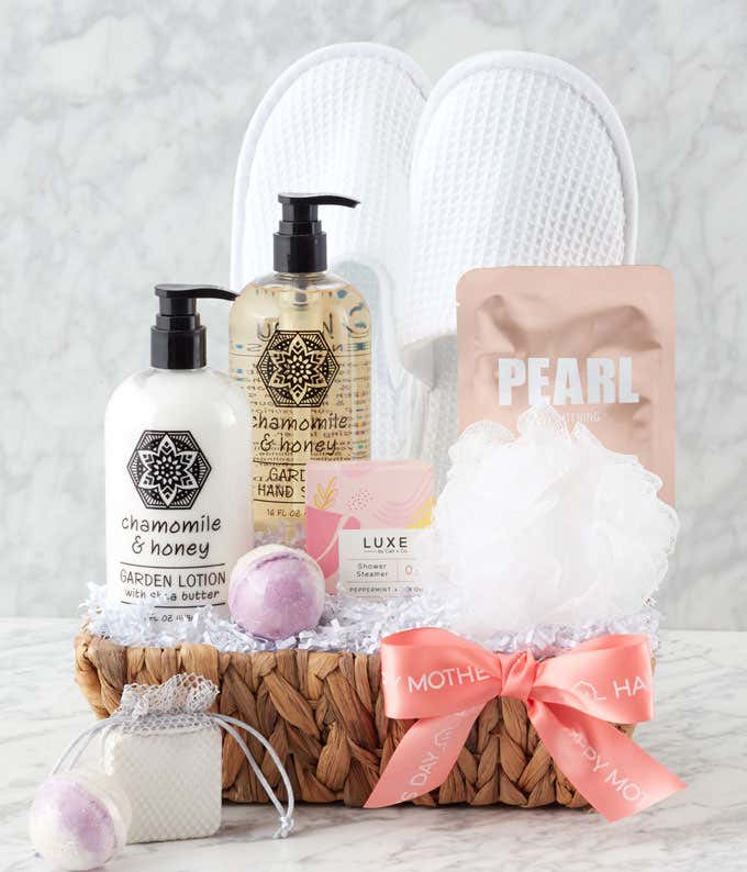 A Mother's Day gift basket filled with Peppermint & Lemon Shower Steamer, Chamomile & Honey Lotion, Body Wash, a small loofah, bath bomb, waffle slippers, and a Pearl Brightening Face Mask, beautifully presented with a ribbon and personalized message.