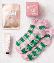 Alternative image 1 for Gift box with a gold pearl brightening face mask, pink magnolia-scented bath bomb, rose-scented hand cream, and a pair of fuzzy pink and green plaid socks with pom-poms, all arranged on white crinkle paper in a pink box.