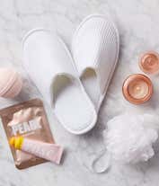 Alternative image 1 for Spa gift set in a beige fabric basket with a white bow, featuring white slippers, bath bombs, pearl face masks, rose-scented lotion, a white loofah, a pink candle, and a pink glass jar, displayed on a marble surface.