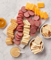 Alternative image 1 for Gift basket with summer sausages, cheeses, crackers, and mustards in a white patterned container with a red bow. A serving board in front displays sliced sausage, cheese, and mustard for dipping.