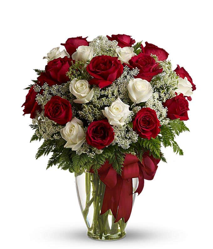 A floral arrangement featuring red roses, white roses, and floral greens in a clear glass vase with a red bow.