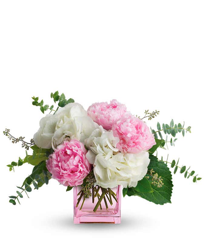 Bouquet of light pink peonies, pale green hydrangeas, and eucalyptus stems arranged in a square pink glass vase.