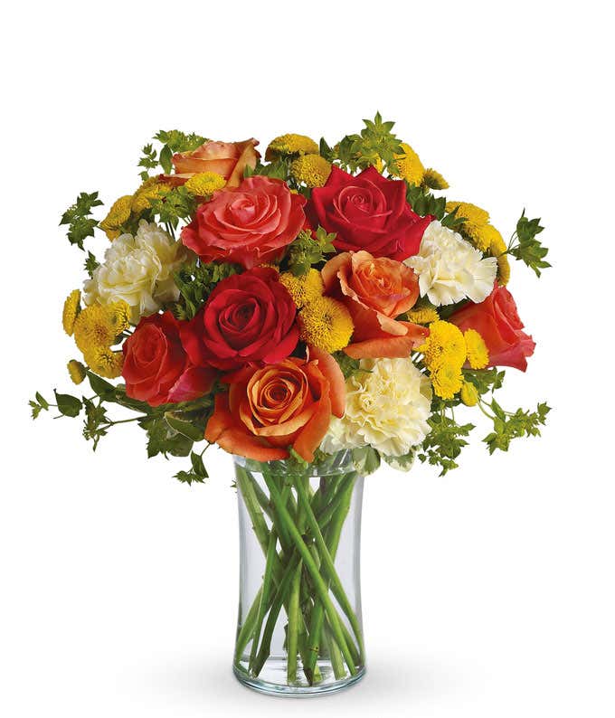 A vibrant floral arrangement featuring orange roses, coral roses, white carnations, and yellow button mums, all beautifully displayed in a glass gathering vase.