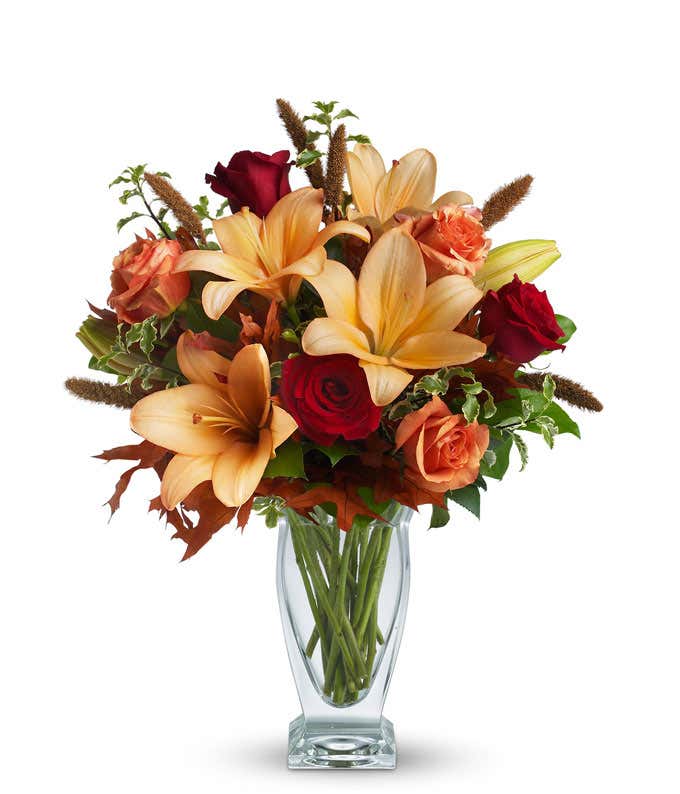 A stunning floral arrangement featuring orange roses, red roses, orange Asiatic lilies, and dried leaves, all elegantly presented in a glass vase.