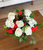 Alternative image 1 for A beautiful floral arrangement with red mini carnations, white roses (deluxe and premium sizes only), white cushion spray chrysanthemums, Douglas fir, variegated holly, noble fir, pinecones, and red berries, displayed in a clear cube vase.