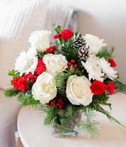 Alternative image 2 for A beautiful floral arrangement with red mini carnations, white roses (deluxe and premium sizes only), white cushion spray chrysanthemums, Douglas fir, variegated holly, noble fir, pinecones, and red berries, displayed in a clear cube vase.