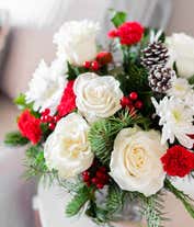 Alternative image 3 for A beautiful floral arrangement with red mini carnations, white roses (deluxe and premium sizes only), white cushion spray chrysanthemums, Douglas fir, variegated holly, noble fir, pinecones, and red berries, displayed in a clear cube vase.