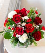 Alternative image 1 for A festive arrangement with red roses (deluxe & premium only), red carnations, white carnations, red mini-carnations, white mini-carnations, seasonal greens, silver ornaments, and pine cones.