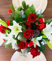 Alternative image 1 for A festive arrangement with red roses, white Asiatic lilies, red mini-carnations, seasonal greens, silver ornaments, and pinecones, beautifully displayed in a clear cylinder vase.