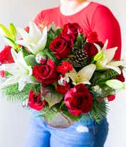 Alternative image 2 for A festive arrangement with red roses, white Asiatic lilies, red mini-carnations, seasonal greens, silver ornaments, and pinecones, beautifully displayed in a clear cylinder vase.
