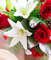 Alternative image 3 for A festive arrangement with red roses, white Asiatic lilies, red mini-carnations, seasonal greens, silver ornaments, and pinecones, beautifully displayed in a clear cylinder vase.