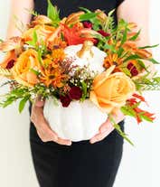 Alternative image 2 for A charming floral arrangement featuring orange roses, orange alstroemeria, maroon mini carnations, yellow mums, and white waxflower, all beautifully arranged in a ceramic pumpkin.