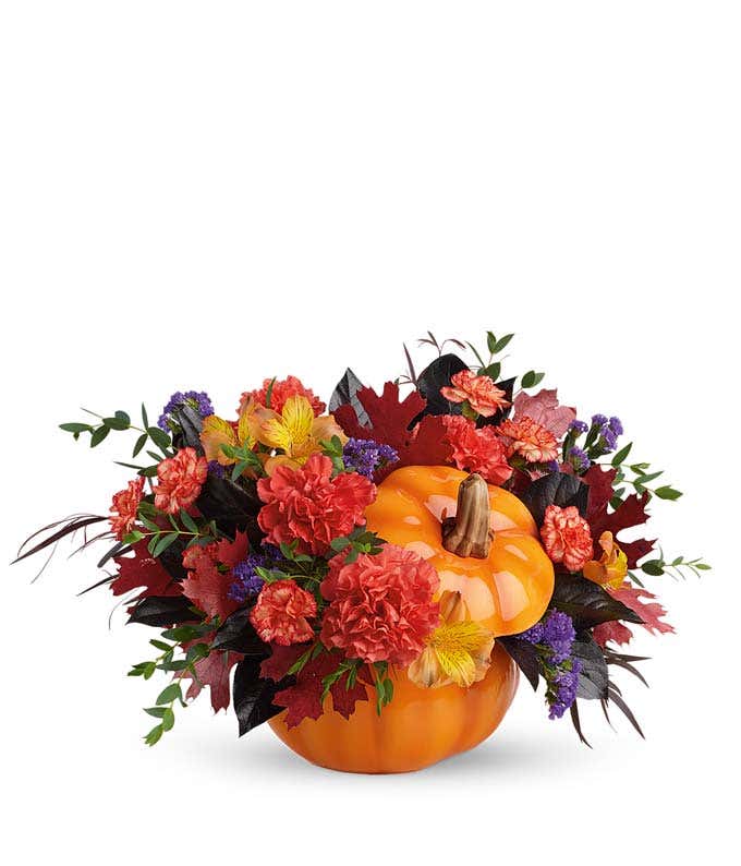A vibrant floral arrangement featuring orange carnations, orange mini carnations, orange Peruvian lilies, purple statice, deluxe and premium bi-color roses, and oak leaves, all beautifully presented in a keepsake container.
