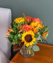 Alternative image 1 for A bright floral arrangement featuring deluxe and premium orange roses, sunflowers, orange safflower, orange Peruvian lilies, and yellow solidago, all beautifully arranged in a cube vase.