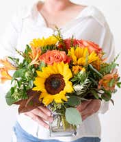 Alternative image 2 for A bright floral arrangement featuring deluxe and premium orange roses, sunflowers, orange safflower, orange Peruvian lilies, and yellow solidago, all beautifully arranged in a cube vase.