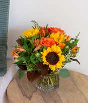 Alternative image 3 for A bright floral arrangement featuring deluxe and premium orange roses, sunflowers, orange safflower, orange Peruvian lilies, and yellow solidago, all beautifully arranged in a cube vase.