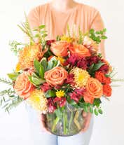 Alternative image 1 for A vibrant floral arrangement featuring orange roses, orange spray roses, red Peruvian lilies, bronze mums, and orange daisies, all elegantly displayed in a clear cylinder vase.