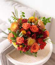 Alternative image 2 for A vibrant floral arrangement featuring orange roses, orange spray roses, red Peruvian lilies, bronze mums, and orange daisies, all elegantly displayed in a clear cylinder vase.