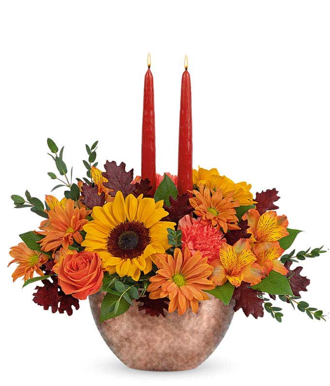  A cheerful floral arrangement featuring sunflowers, orange carnations, bronze daisies, orange alstroemeria, and seasonal greenery, complemented by taper candles, all beautifully presented in a keepsake bowl.