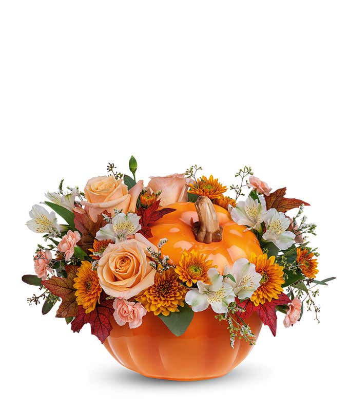 Floral arrangement of peach roses, white alstroemeria, miniature peach carnations, and bronze cushion spray chrysanthemums with greenery, displayed in a bright orange pumpkin-shaped container.