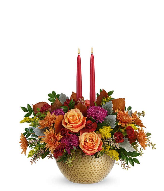 A rich floral arrangement featuring orange roses, maroon miniature carnations, rust and purple cushion spray chrysanthemums, solidago, brown copper beech, dusty miller, seeded eucalyptus, and huckleberry, complemented by burgundy candles, all beautifully 