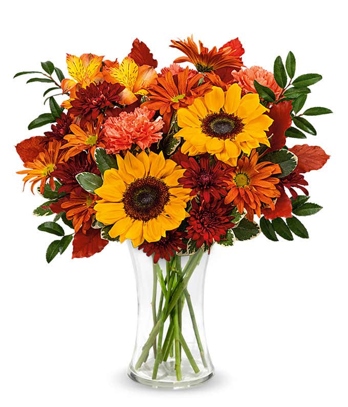 A cheerful floral arrangement featuring yellow sunflowers, orange carnations, orange alstroemeria, bronze daisy spray chrysanthemums, bronze cushion spray chrysanthemums, huckleberry, variegated pittosporum, and burgundy copper beech, all beautifully disp