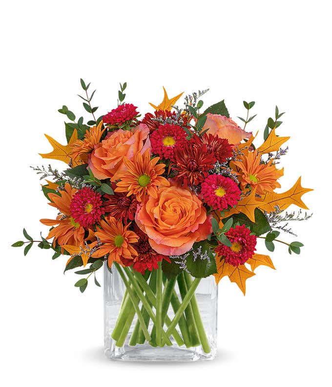 A vibrant floral arrangement featuring orange roses, red Matsumoto asters, bronze cushion spray chrysanthemums, bronze daisy spray chrysanthemums, purple limonium, parvifolia eucalyptus, and yellow oak leaves, all elegantly arranged in a bronze vase.